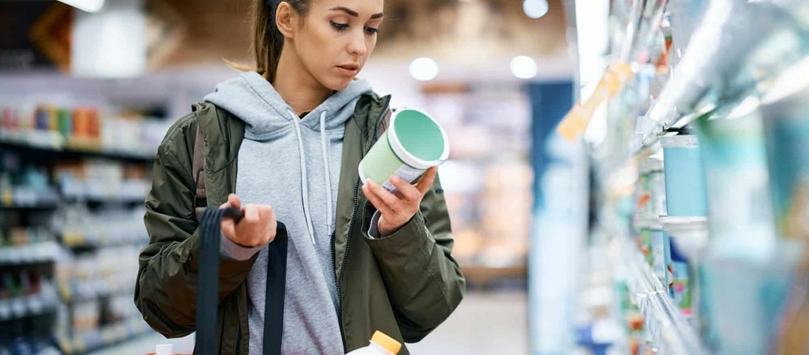 Young woman reading nutrition label while buying diary product in supermarket.